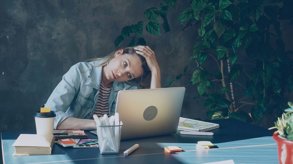 Jovem adulta cansada à secretária com computador, representando burnout, exaustão emocional e sobrecarga.