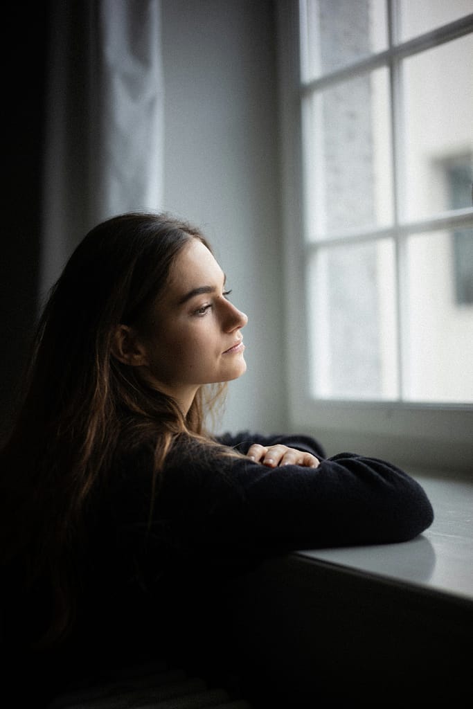 Mulher encostada à janela a olhar para fora com expressão pensativa, num ambiente calmo e iluminado por luz natural representando luto e perda